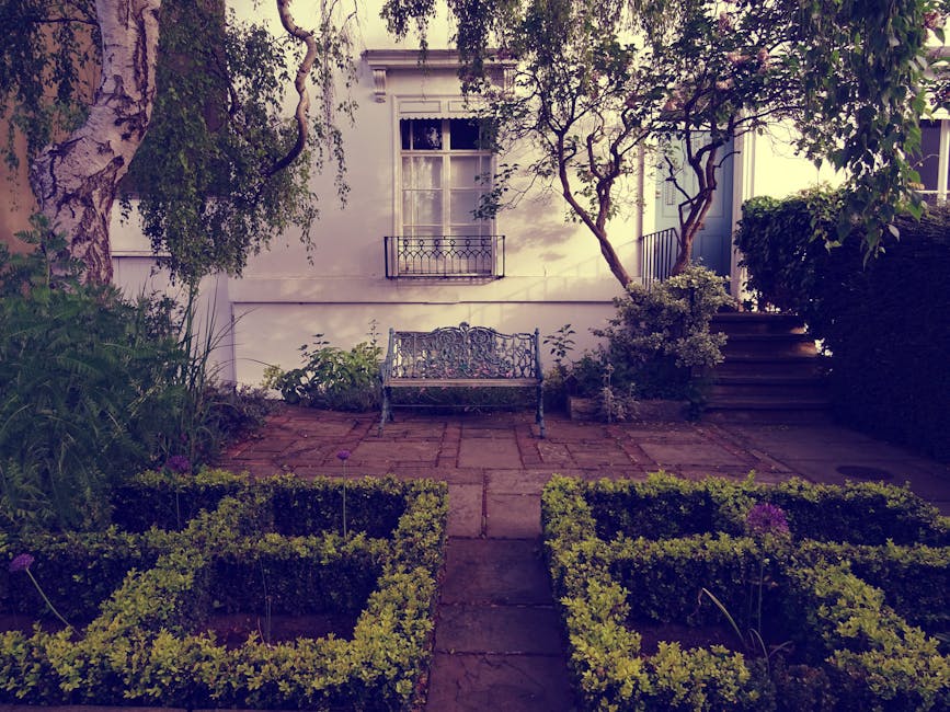 A small front garden featuring a paved pathway leading to a decorative metal garden bench positioned against a white exterior wall with a window and a small balcony. The garden is surrounded by neatly
