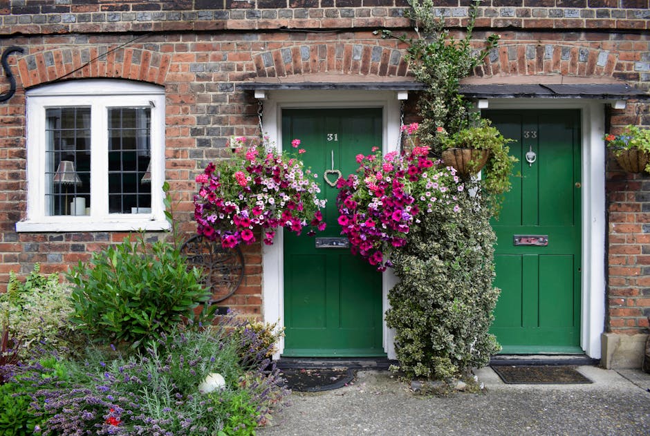 A small front garden featuring a paved pathway leading to a decorative metal garden bench positioned against a white exterior wall with a window and a small balcony. The garden is surrounded by neatly